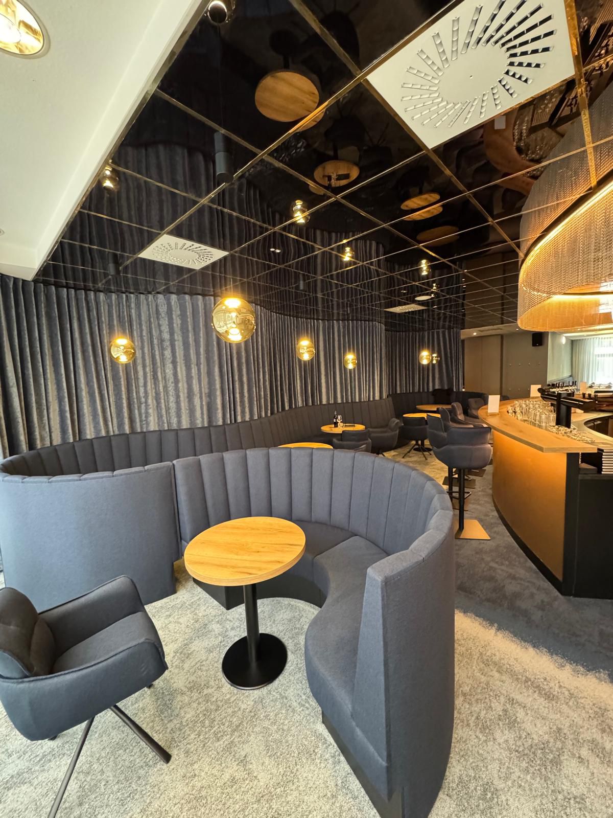Eine elegante Lounge mit grauen Sitznischen, runden Holztischen und goldenen Lampen